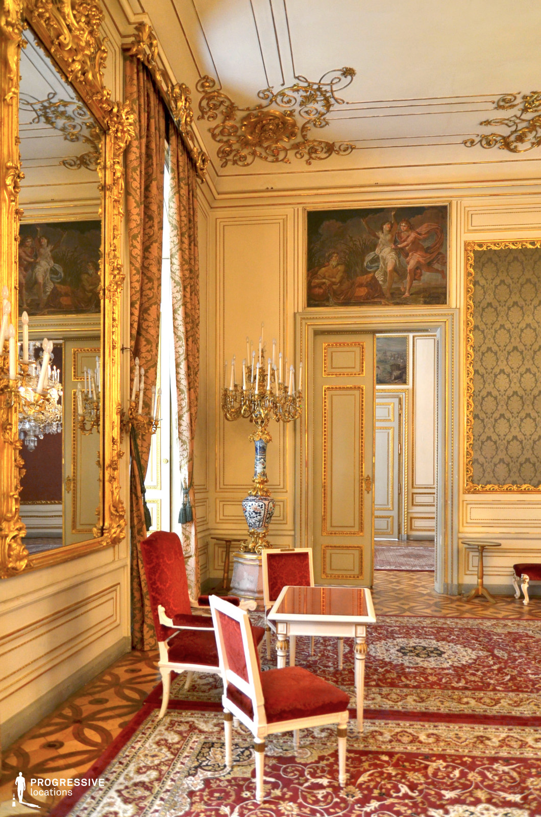 This opulent Rococo-style salon features intricate gold moldings, grand mirrors, and richly upholstered furniture that exude aristocratic elegance. The pale cream walls are accented with gilded carvings and classical frescoes, adding depth and refinement to the space. A plush, patterned red carpet complements the velvet-upholstered chairs and delicate white-and-gold furniture, reinforcing the regal aesthetic. A candelabra-style floor lamp and elegant drapery contribute to the room’s historical ambiance. This setting is ideal for period dramas, royal narratives, and high-society intrigue, offering a richly detailed and visually sophisticated backdrop.
