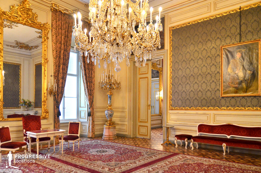 This opulent salon exudes classical elegance with its intricate gilded moldings, large crystal chandelier, and richly patterned wallpaper framed by gold accents. The high ceilings and tall windows draped in heavy brocade curtains enhance the grandeur, while the parquet flooring is softened by luxurious Persian rugs. An oversized gilded mirror reflects the lavish interior, adding depth and sophistication. The refined furniture, including velvet-upholstered seating and carved wooden tables, contributes to the aristocratic atmosphere. This setting is ideal for period dramas, historical romances, or regal political intrigues, offering a sophisticated and richly detailed backdrop for high-society gatherings, diplomatic meetings, or intimate aristocratic encounters.