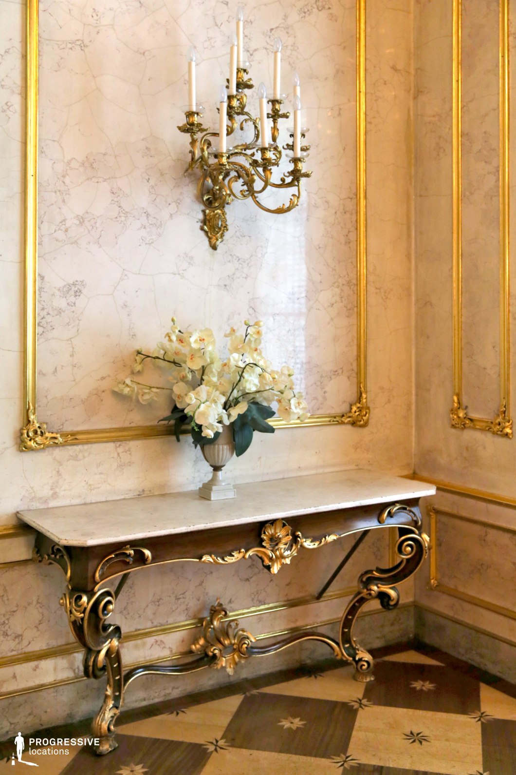 This refined Rococo-style interior features an elegant marble console table with intricate gilded detailing, gracefully curved legs, and a polished stone top. Above it, an opulent gold wall sconce with candle-like lights enhances the classical ambiance. The marble wall, framed with delicate gilded moldings, adds a luxurious and sophisticated touch. The parquet flooring, adorned with star-like inlays, complements the richness of the decor. This location is ideal for period dramas, aristocratic settings, or high-society film sequences, offering a backdrop of elegance, refinement, and historical grandeur.