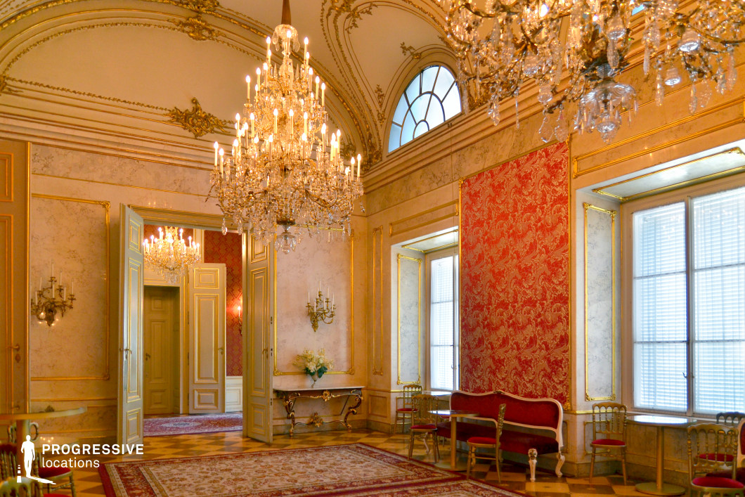 This elegant hall exudes aristocratic refinement with its gilded moldings, high ceilings, and intricate marble wall detailing. The grand crystal chandeliers, suspended from ornately decorated ceilings, add a dazzling focal point, while the tall arched windows and mirrored surfaces enhance the sense of space and opulence. Rich red damask wall panels contrast with the light-colored stone and gold trim, creating a warm yet regal ambiance. The open double doors provide depth and continuity between adjoining rooms, making this an ideal filming location for period dramas, royal court scenes, and historical narratives. The soft, natural light filtering through the windows, combined with the lavish decor, offers a visually striking setting for formal gatherings, diplomatic meetings, or aristocratic intrigue.