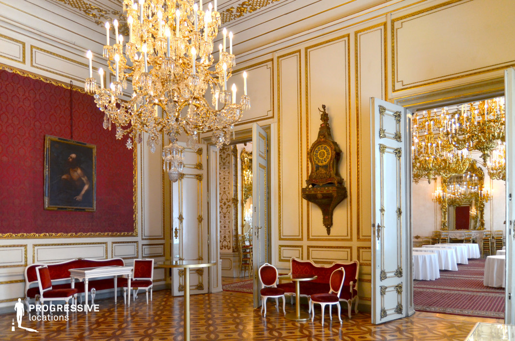 This opulent salon showcases a rich combination of deep red damask walls, gilded moldings, and elegant white paneling, creating a regal and sophisticated ambiance. A grand crystal chandelier hangs from the ceiling, casting a warm glow over the intricately patterned parquet flooring. The space is furnished with plush red velvet seating and delicate white-framed tables, enhancing its aristocratic charm. Through the open double doors, an adjoining ballroom with ornate chandeliers and mirrors adds depth and grandeur. This setting is ideal for filming period dramas, royal intrigues, or historical romances, offering a lavish backdrop filled with refinement and elegance.