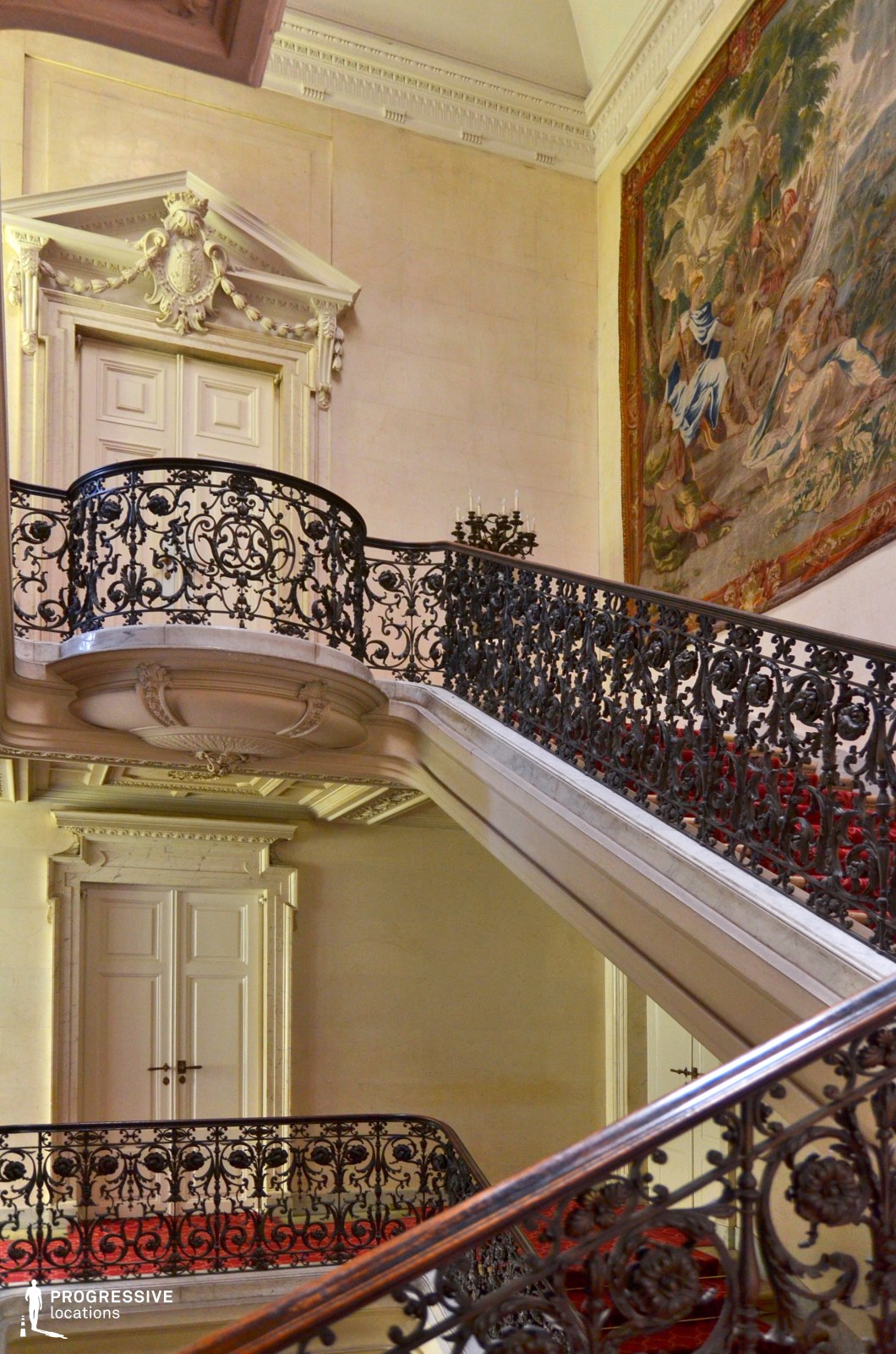 This grand staircase embodies Baroque elegance with its intricate wrought-iron railings, richly decorated moldings, and a striking wall tapestry depicting a classical scene. The pale stone walls contrast beautifully with the deep red carpeting, adding a regal atmosphere. The upper landing features a prominent doorway framed by an elaborate pediment adorned with sculptural details, reinforcing the space’s aristocratic character. The gracefully curved staircase and ornamental balcony create a dynamic composition, making this an ideal setting for historical dramas, period films, or luxurious high-society intrigue.
