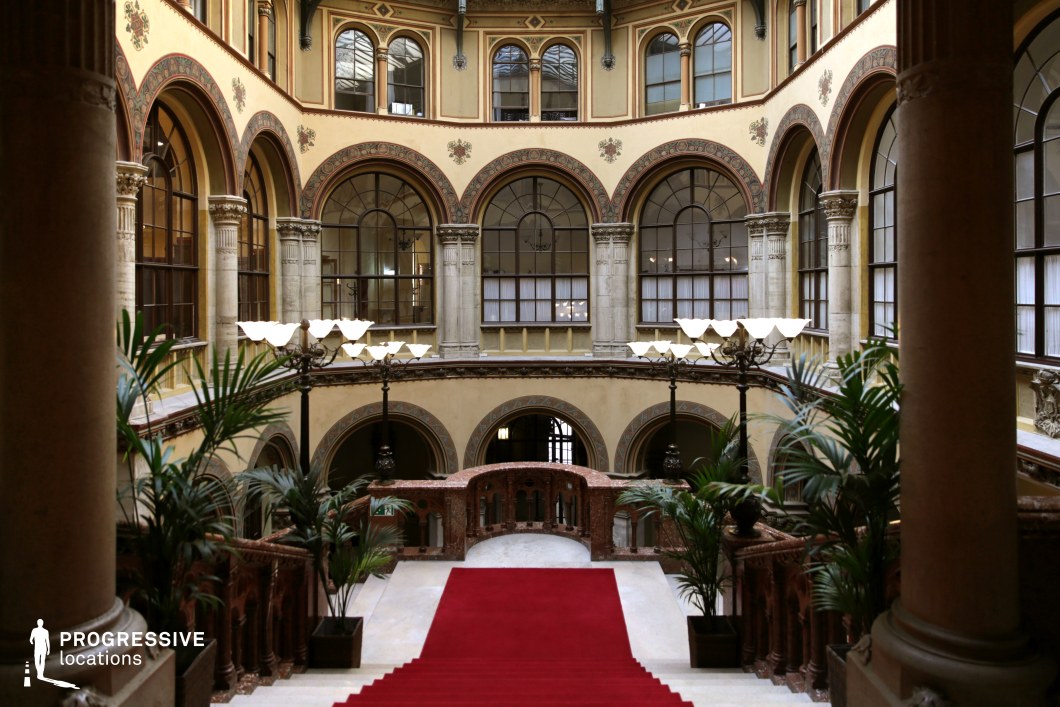 This opulent interior features a sweeping staircase covered in a deep red carpet, framed by intricate stone balustrades and lush decorative plants. The surrounding space is defined by tall, rounded archways adorned with ornate frescoes, classical columns, and large windows that allow soft natural light to filter in. Elegant wrought-iron lampposts add a regal touch, enhancing the sense of luxury and refinement. The richly detailed architecture and grand symmetry make this location ideal for period dramas, aristocratic settings, or high-society intrigue, providing a visually stunning backdrop for sophisticated, cinematic storytelling.