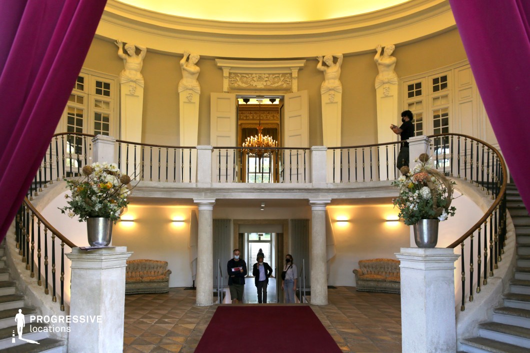 This opulent neoclassical entrance hall features a symmetrical double staircase with elegant wrought-iron railings and a polished stone finish, leading to a semi-circular mezzanine. The grand architectural design is accentuated by towering caryatid sculptures, which add a classical and majestic touch to the space. A deep red carpet extends towards an intricately carved doorway, through which a warmly lit chandelier glows, enhancing the regal ambiance. The neutral-toned walls and checkered stone flooring contrast with rich magenta curtains, framing the scene with a theatrical elegance. This setting is ideal for period dramas, high-society gatherings, or suspenseful thriller scenes, providing a luxurious yet slightly mysterious atmosphere for film productions.
