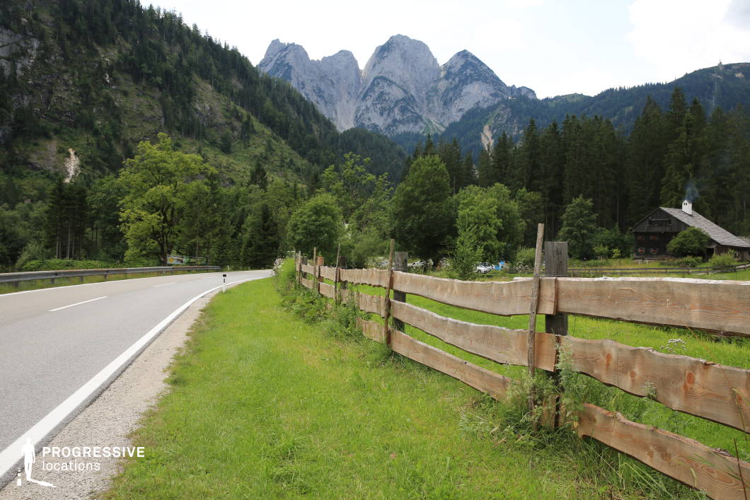 This location captures a gently curving countryside road bordered by a raw-cut wooden fence that divides the asphalt from a green pastureland. The fence, with its uneven planks and natural finish, adds a rustic texture to the composition and leads the eye toward a dark timber house partially hidden by dense pine forest. Towering behind the treeline, a rugged mountain range dominates the background with steep rock faces and pointed peaks, reinforcing the sense of seclusion and natural grandeur. The color palette consists of rich greens, muted browns, and greys, evoking a grounded, organic mood. The camera angle, taken from road level, emphasizes the linear progression of the fence and road into the deep natural landscape. This location is highly adaptable for film productions requiring a serene rural setting, and is well-suited for drama, historical narratives, or travel-related scenes where connection to nature and traditional architecture are key visual elements.