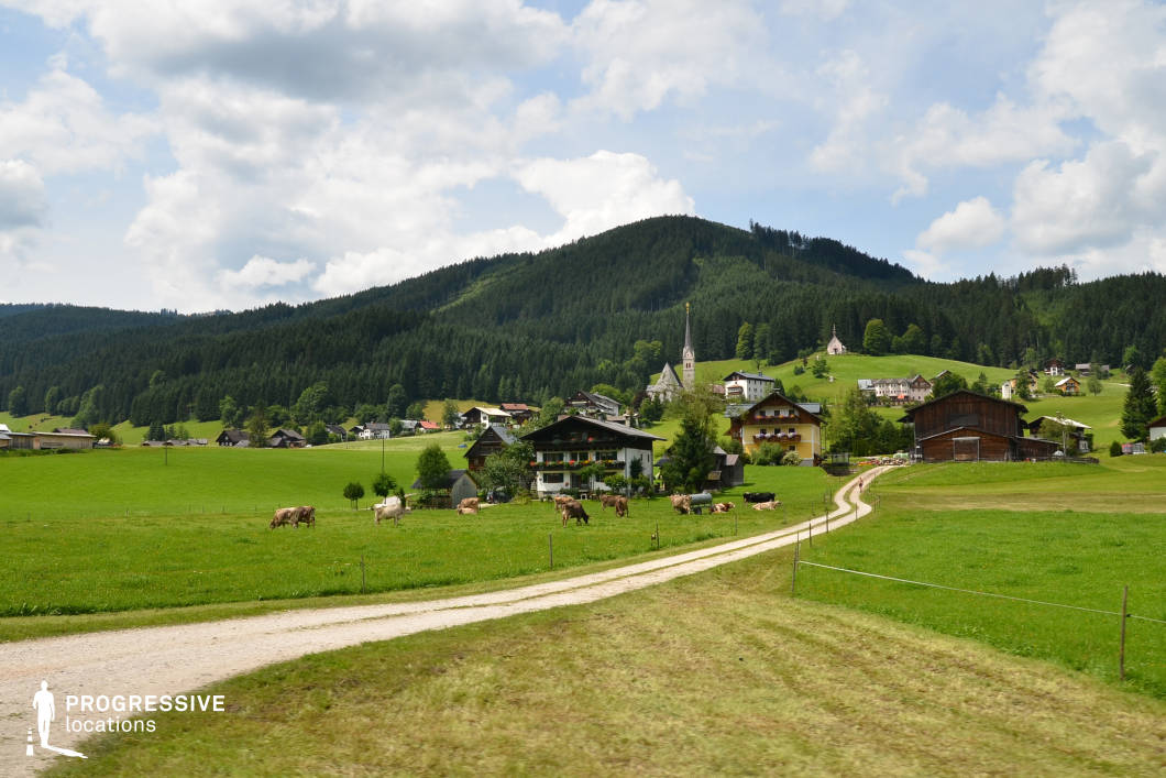 This location features a traditional alpine village nestled in a wide, open valley bordered by forested hillsides. The composition highlights a gravel path leading toward clusters of chalet-style buildings with wooden balconies and sloped roofs, surrounded by vibrant green fields and gently rolling terrain. The architecture is cohesive, with a mix of white facades and natural timber elements, contributing to a harmonious rural aesthetic. A church with a pointed steeple anchors the village visually, while the forested backdrop provides a sense of enclosure and depth. The scene is structured with clear foreground, middle ground, and background layers, making it highly versatile for wide establishing shots. This location is ideal for genres such as drama, romance, or period pieces, and supports scenes that benefit from idyllic, pastoral settings with traditional European character.