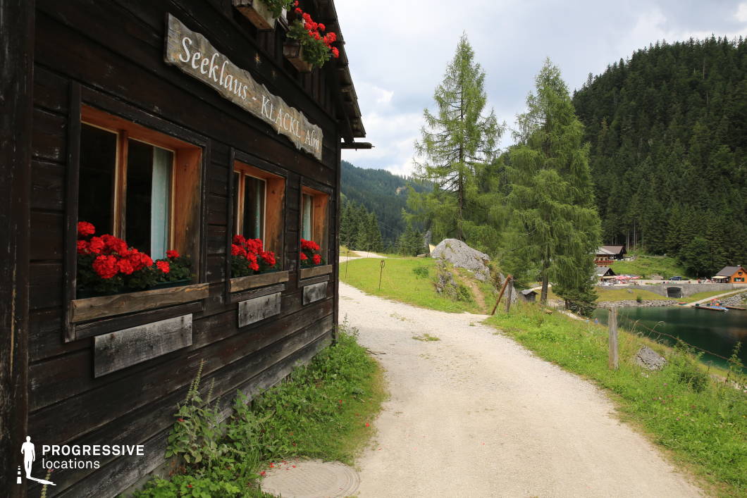 This charming location features a dark wooden cabin with a weathered exterior and vibrant flower boxes beneath each window, adding strong color contrast and visual warmth to the scene. The narrow gravel path beside the building curves gently through a green, forested landscape and leads toward a lakeside area with small structures and conifer-covered slopes in the background. The use of natural materials, such as timber and stone, blends harmoniously with the alpine environment, while the rustic architecture provides a sense of tradition and simplicity. The composition creates a visual narrative between built and natural elements, supported by the vertical presence of tall trees and the reflective surface of the water. This location is ideal for period dramas, documentary segments, or commercial shoots focused on alpine tourism or pastoral tranquility.
