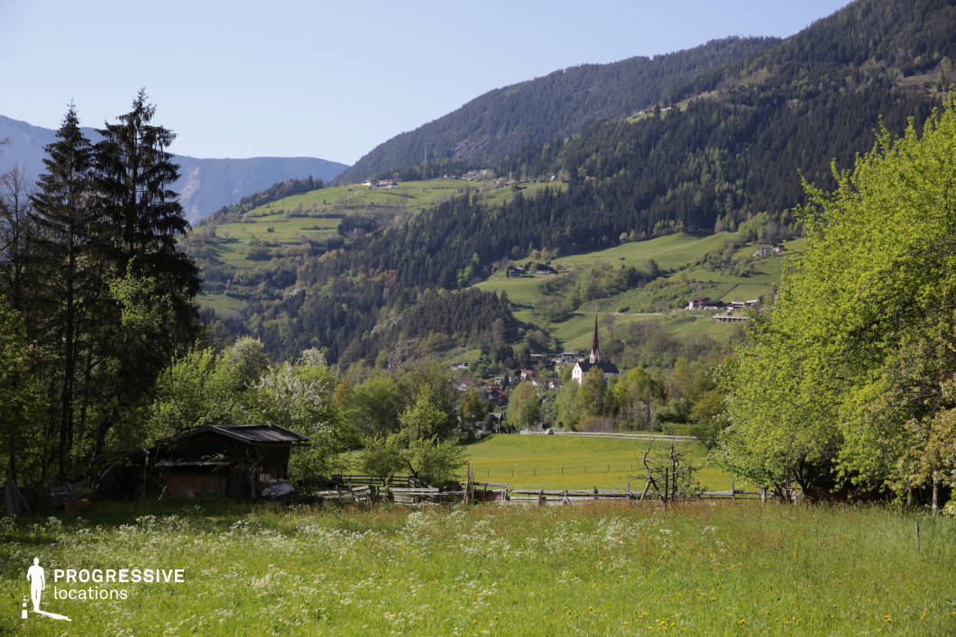 This location showcases a wide alpine valley characterized by rolling green meadows, a patchwork of forested hillsides, and terraced pastures extending up the mountainsides. The image is anchored by a white village church with a tall, pointed spire, positioned centrally in the midground, offering a strong vertical accent against the horizontal layering of the landscape. The foreground features a rustic wooden structure and a wooden fence, which reinforce the pastoral, traditional atmosphere. The dominant color palette consists of rich greens, natural wood tones, and soft earth hues, evoking a tranquil and unspoiled rural charm. The varying elevation creates a dynamic visual rhythm, with soft hills leading to steeper, pine-covered slopes, while the perspective provides a deep sense of spatial continuity from foreground to horizon. This setting is highly suitable for filming period dramas, nature documentaries, or romantic and introspective scenes, offering both visual serenity and authentic rural textures.