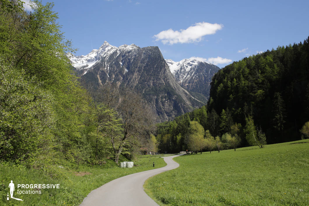 This location presents a gently winding paved road nestled within a wide, open mountain valley, flanked by dense forests and set against the dramatic backdrop of rugged, snow-capped peaks. The expansive green meadow on the right and the layered woodland on the left create a balanced composition that contrasts vertical and horizontal elements. The towering rock faces with their textured geological formations add a raw, natural monumentality to the scene. The dominant color palette—vivid greens, deep grays, and whites—conveys a clean, serene atmosphere with strong natural contrasts. The open landscape, combined with the deep perspective and central leading line of the road, makes this location ideal for cinematic wide shots. It is particularly well-suited for adventure films, road movies, or any genre requiring a striking natural backdrop or a sense of journey and scale.