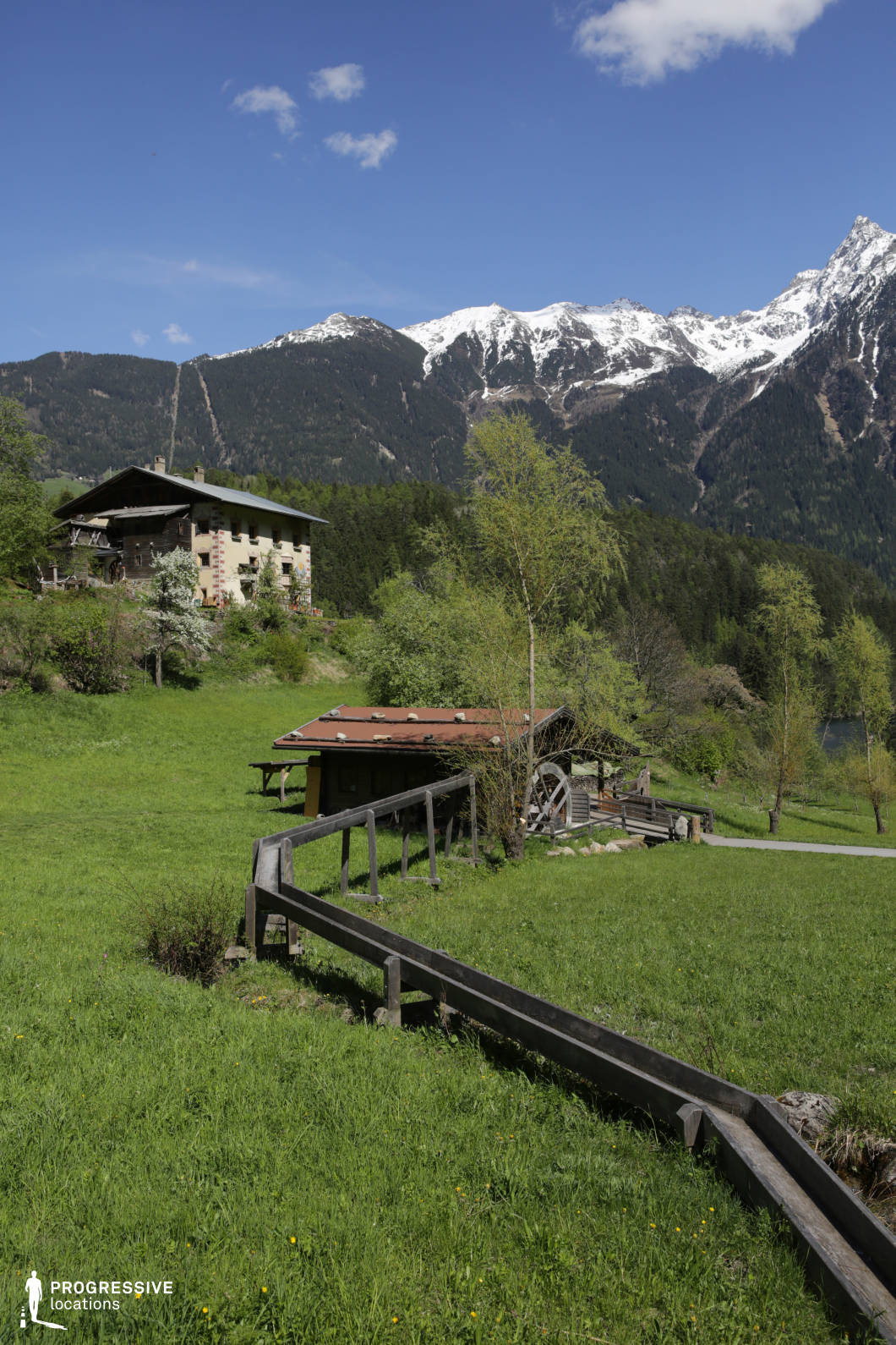This rural location presents a traditional watermill structure and rustic buildings set within a gently sloping grassy meadow, framed by dense forest and snow-capped mountain peaks in the background. A wooden aqueduct runs diagonally across the foreground, visually linking the elements of the composition and emphasizing the historical utility of the space. The architecture features natural wood and plaster textures, blending harmoniously with the green surroundings and reinforcing the pastoral atmosphere. The surrounding forest forms a natural boundary at the base of the mountains, while the jagged peaks rise sharply, providing a dramatic contrast to the softness of the foreground. The vertical format of the image captures the full elevation of the landscape from the meadow to the summits. This setting is highly versatile for filming, ideal for period pieces, countryside scenes, or nature-centric productions in genres such as historical drama, fantasy, or environmental documentaries, offering authenticity, visual layering, and rich textural detail.