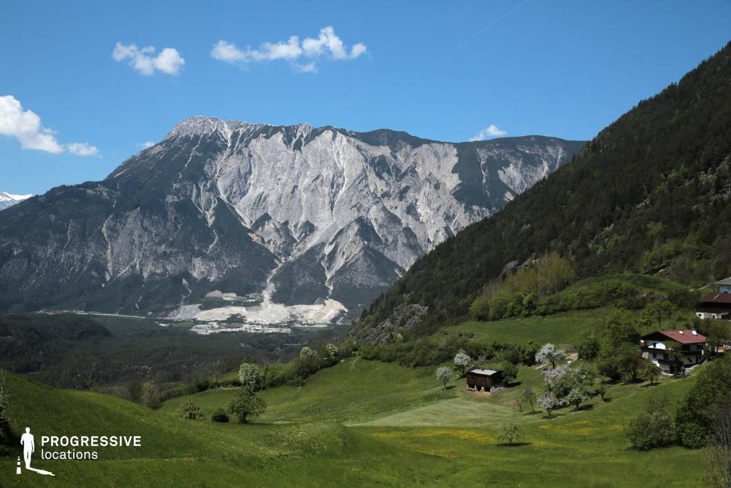 This location features a dramatic natural landscape characterized by a vast alpine meadow in the foreground and a monumental, jagged mountain range dominating the background. The sloping green terrain is interspersed with scattered trees and traditional wooden buildings, set against a steep forested hillside that adds vertical contrast. The mountain’s exposed rock face displays sharp, angular formations in varying shades of gray, contributing to a striking visual tension between natural tranquility and raw geological power. The open, expansive setting and distinct layering of foreground and background elements provide excellent depth and compositional clarity, making it highly suitable for landscape-focused scenes, establishing shots, or visually poetic sequences in genres such as drama, arthouse cinema, or documentaries.