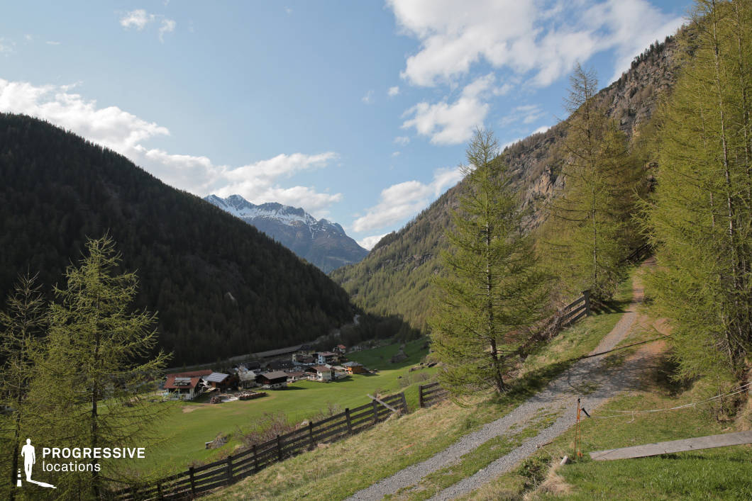 This wide-angle view captures a peaceful alpine valley framed by densely forested mountain slopes and snow-capped peaks in the distance. A narrow gravel path curves gently along the hillside in the foreground, bordered by sparse larch trees and rustic wooden fencing. Below, a small village with pitched-roof buildings is nestled in a green clearing, providing scale and grounding within the vast natural surroundings. The composition offers layered depth from foreground to background, supported by a harmonious blend of earthy and cool tones. This location is well-suited for cinematic sequences requiring a tranquil yet expansive rural backdrop, ideal for drama, period pieces, or nature-focused documentaries.