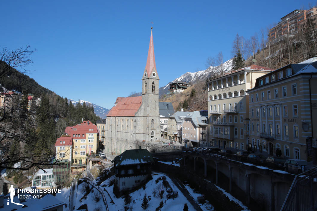 This elevated townscape features a central Gothic-style stone church with a tall, pointed spire that serves as the dominant visual anchor, surrounded by multi-story historic buildings with pastel facades and classic architectural detailing. The narrow, winding layout of the streets and the terraced buildings cascading down the hillside emphasize the verticality and density of the town, which is nestled within a mountainous landscape. The stone textures of the church and surrounding walls contrast with the smoother plastered facades of the neighboring structures, creating a visually engaging blend of materials. The perspective provides a dynamic view from an upper vantage point, capturing the full scale of the valley settlement. This location is ideal for filming scenes set in a European mountain town and lends itself well to genres such as drama, historical fiction, and mystery due to its layered composition and atmospheric architectural setting.
