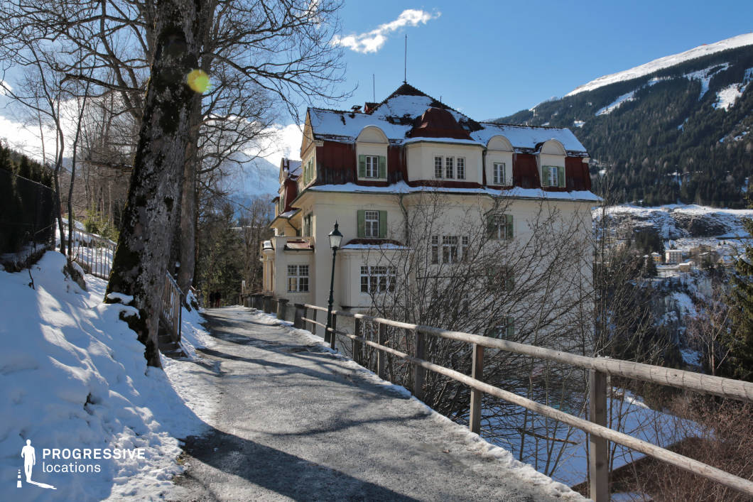 This image captures a narrow, gently winding hillside pathway flanked by a rustic wooden railing on one side and mature, leafless trees on the other, creating a visually balanced composition. The central architectural focus is a grand, historic villa with cream-colored facades and contrasting dark red mansard roofs, featuring arched dormer windows and green shutters. The building’s textured walls and classical proportions evoke early 20th-century alpine architecture, and its elevated position provides a scenic backdrop of snow-covered slopes and densely forested mountains. The path’s gravel surface and moderate width enhance the location’s authenticity, while the surrounding topography and railing lines offer strong linear perspective ideal for dynamic framing. This location is ideal for filming period dramas, psychological thrillers, or introspective scenes, particularly those requiring a visually rich, secluded mountain town ambiance.