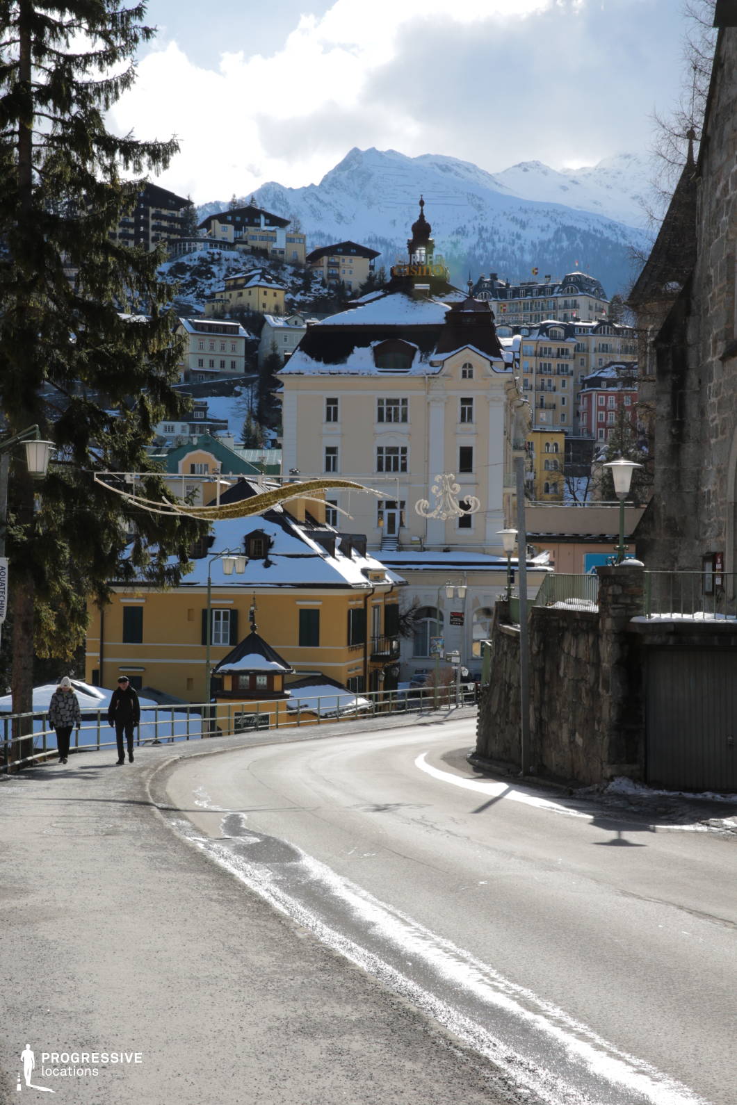 This location features a gently curving asphalt road winding through a historic alpine townscape, framed by traditional multi-story buildings with varied facades and classical architectural details. The immediate foreground includes a stone retaining wall and metal railing, which contribute to a narrow, slightly enclosed spatial feel, while the background expands into a hillside densely layered with pastel-toned and earth-colored structures. Architectural elements such as mansard roofs, arched windows, and ornate detailing evoke a Central European aesthetic. The vertical layering of buildings rising up the mountain slope adds depth and visual rhythm, enhanced by the contrasting textures of stone, plaster, and wood. This setting is particularly well-suited for filming atmospheric urban sequences, character-driven narratives, or period scenes, offering a visually rich, cohesive environment that supports drama, mystery, or historical genres.