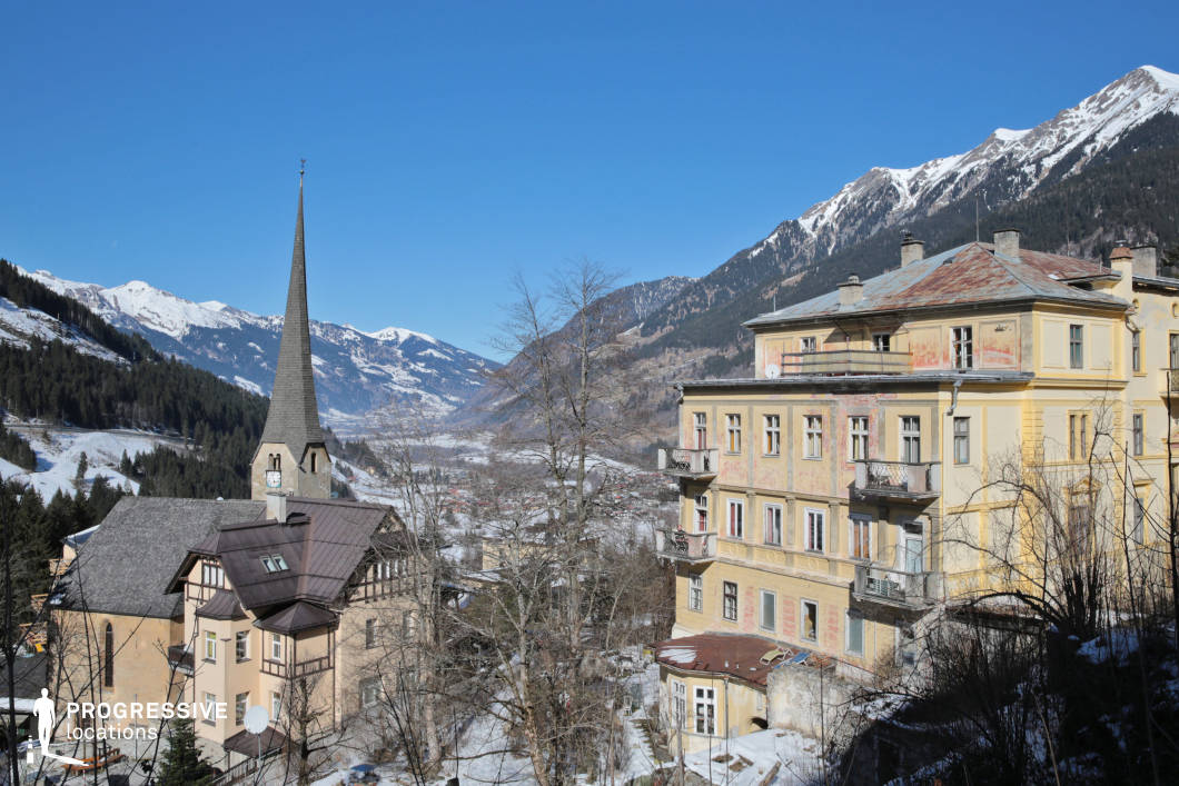 This panoramic location captures a picturesque alpine town nestled in a mountainous valley, characterized by a striking blend of historic European architecture and dramatic natural topography. Dominating the left side of the composition is a pointed church spire rising above a multi-gabled building with stone and timber features, while the right side showcases a four-story, slightly weathered neoclassical-style residential structure with ornate balconies and faded pastel facades. The visual rhythm of the architecture is enhanced by the linear perspective of the valley and the steep, snow-covered mountain slopes in the background. The color palette is defined by muted yellows, browns, and soft greys contrasted against the deep greens of the coniferous forest and the rocky white of the peaks. The framing is slightly elevated, offering a sweeping view that emphasizes vertical contrasts and depth. This setting is ideal for filming projects requiring a distinctive European mountain village aesthetic, suitable for period pieces, atmospheric dramas, or mystery genres, and is especially fitting for establishing shots or narrative transitions involving isolated or remote environments.