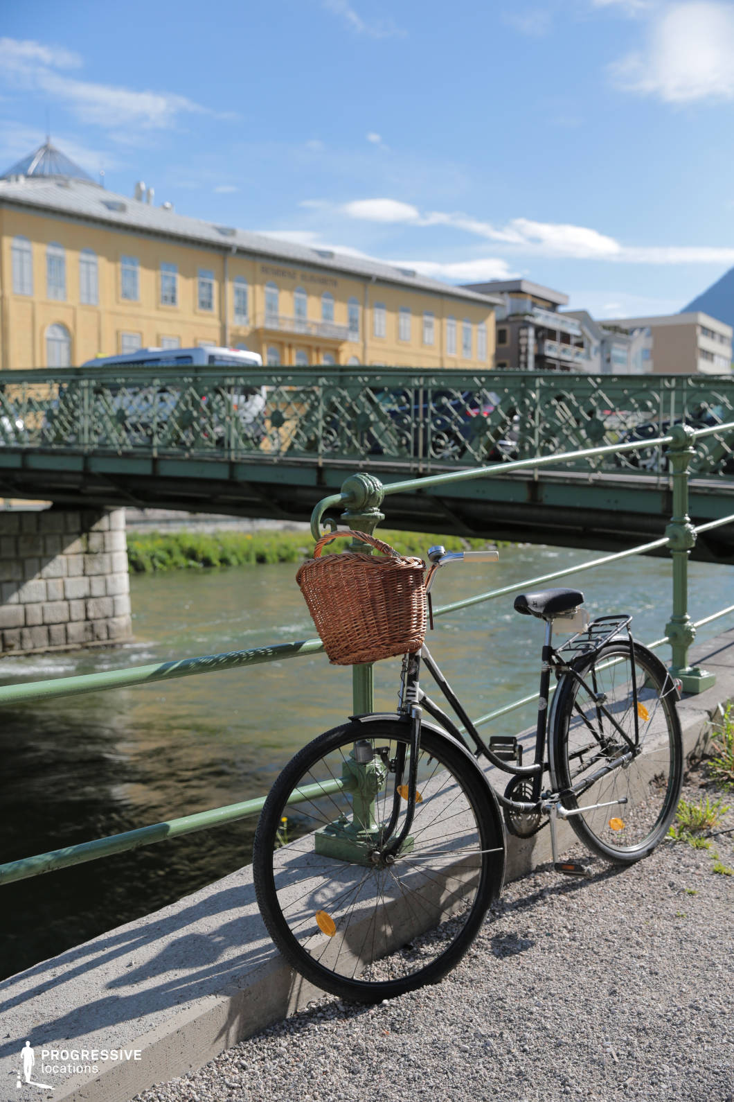 This location features a decorative green iron bridge crossing over a gently flowing river, with detailed latticework and classical stone supports, contributing to a refined historical atmosphere. In the foreground, a black bicycle with a woven basket leans against a matching green railing, echoing the ornate iron patterns of the bridge behind it. The background includes a uniform, pale yellow neoclassical-style building with symmetrical windows and a modest cornice, reinforcing the sense of a well-preserved European townscape. The color palette is dominated by muted greens, yellows, and grays, creating a calm and timeless visual impression. The perspective, captured at street level with a clear side view of the bridge, emphasizes linear elements and depth, making this an ideal location for filming urban riverside scenes, travel montages, or period settings. It suits genres such as romantic drama, historical fiction, or lighthearted European travel narratives.