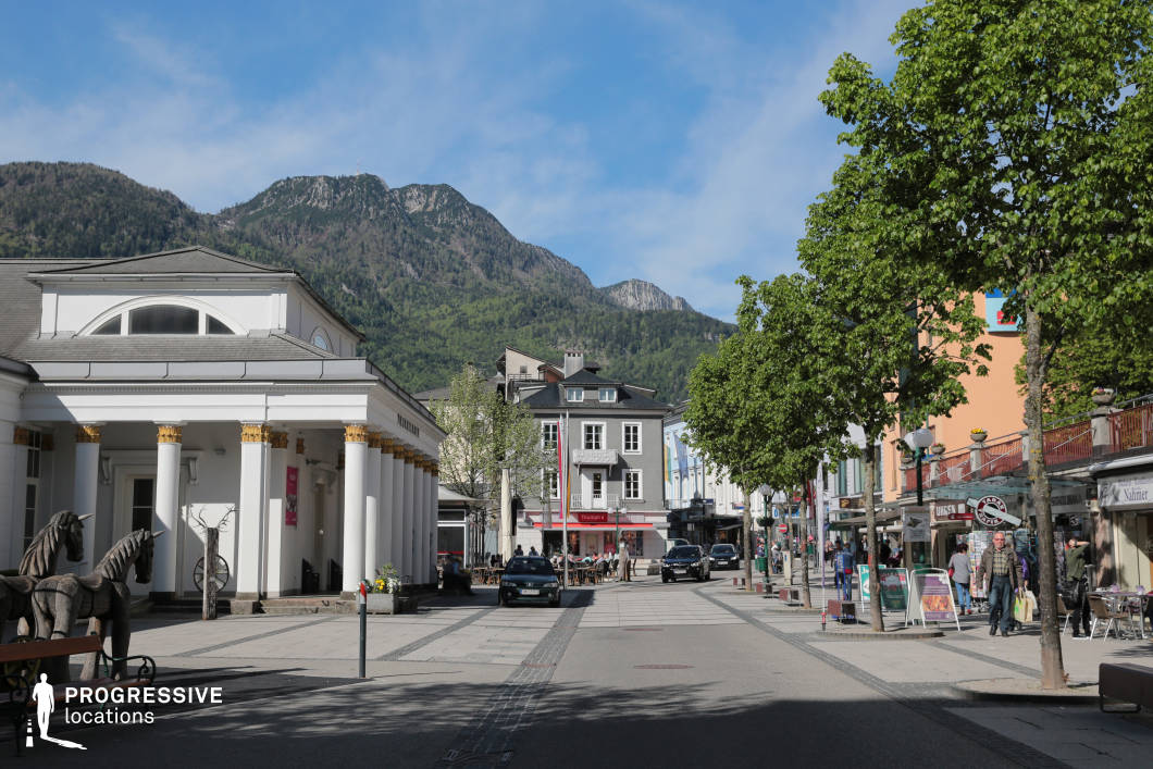 This urban location presents a visually balanced and refined town square framed by a neoclassical colonnaded building on the left, distinguished by its white facade and gold-accented capitals, which convey a formal, almost ceremonial character. The pedestrian-friendly stone-paved avenue stretches centrally through the scene, flanked by mid-rise buildings in a blend of pastel and neutral tones, embodying a small-town European aesthetic. The architecture along the right side exhibits a more casual commercial function, with street-level shops, signage, and an accessible layout. Towering, forested mountains rise in the background, forming a striking natural contrast to the orderly geometry of the town, enhancing the depth of the composition. This setting is ideal for film productions requiring a picturesque small-town European atmosphere, and it suits genres such as drama, romance, or historical narratives. The square's symmetry and distinct architectural elements support wide establishing shots, dialogue scenes, or transitional sequences.