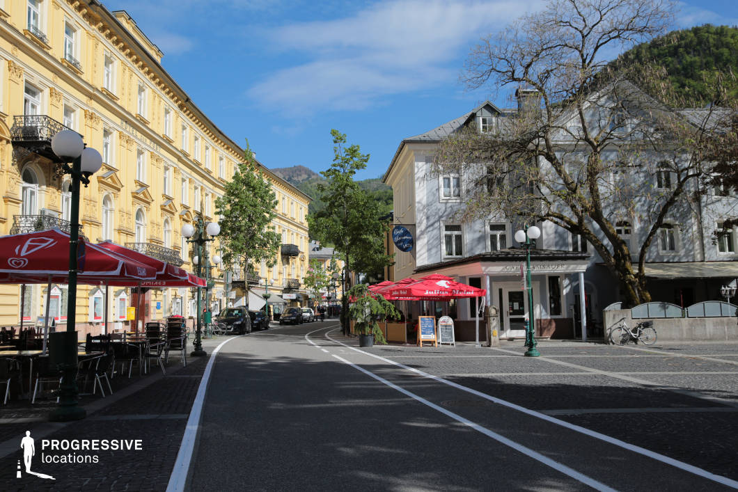 This location presents a gracefully curving urban street flanked by architecturally rich buildings, including a grand, yellow neoclassical facade on the left with ornate window frames and wrought-iron balconies, and a more subdued, traditional grey structure on the right. The street is paved with a combination of smooth asphalt and cobblestones along the pedestrian zones, enhancing its visual texture and historical character. Large, vintage-style streetlamps and evenly spaced trees contribute to the uniform rhythm of the streetscape. Café terraces with red awnings line both sides, suggesting hospitality and leisure as part of the street’s function. The mountain backdrop and visible elevation shifts add depth and context to the environment. This location is ideal for filming elegant small-town settings, European cityscapes, or sophisticated street scenes for genres such as drama, romance, or period pieces.