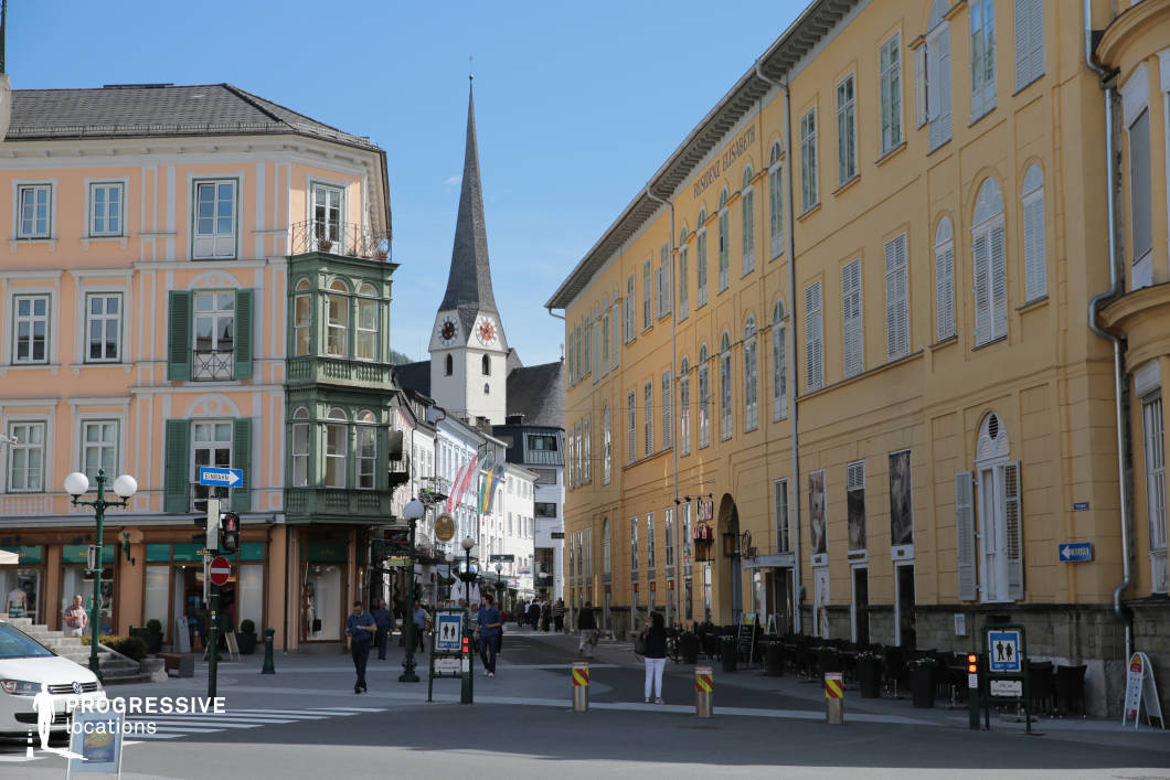 This urban location features a harmonious ensemble of classical European architecture, dominated by pastel and warm yellow façades with ornate window frames and shutter details. A distinctive pointed church tower with a clock face anchors the background, creating a strong vertical focal point and enhancing the town’s historical character. The buildings lining the street exhibit uniformity in height, typically three to four stories, fostering a visually balanced streetscape. Materials such as plastered masonry, wooden shutters, and slate roofing establish a traditional yet well-maintained aesthetic. The street is moderately narrow but open enough to allow clear visibility into the depth of the urban fabric, aided by the perspective of the slight bend. The juxtaposition of arcaded ground-floor commercial units with upper residential or institutional spaces adds functional layering. This setting is ideal for filming romantic dramas, period pieces, or character-driven narratives, especially those requiring a charming, old-world European backdrop. It is suitable for dialogue scenes, walking sequences, and atmospheric establishing shots.
