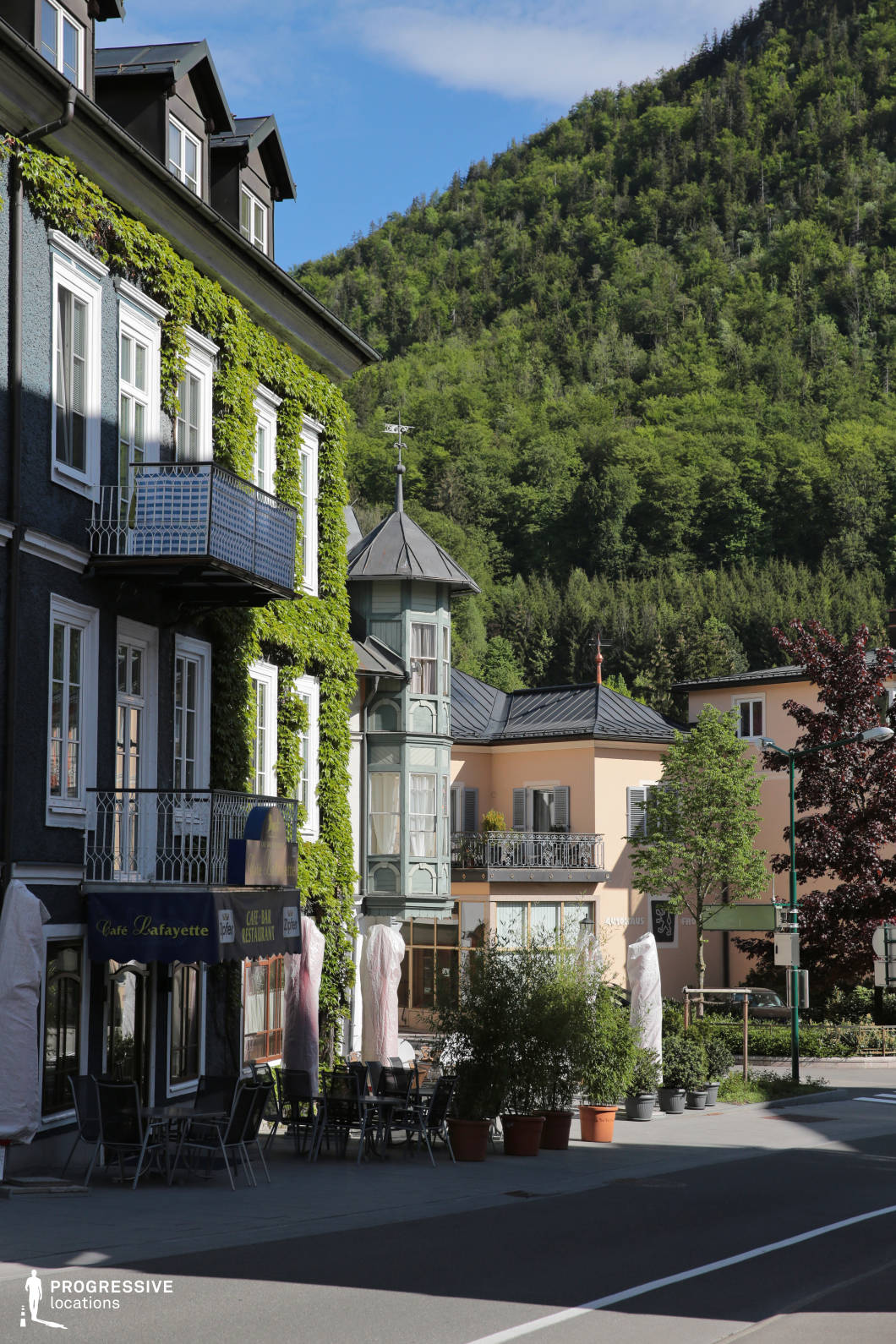 This location features a charming urban streetscape nestled at the foot of a densely forested hillside. The left side of the image is dominated by a multi-story building with dark-colored stucco walls, large white-framed windows, wrought-iron balconies, and a facade partially covered in neatly trimmed climbing vegetation. A standout architectural detail is the narrow, turret-like bay window structure with a pointed roof, adding a fairytale-like quality to the setting. Opposite, pastel-toned buildings with classical window shutters and balconies create a harmonious contrast in color and texture. The paved pedestrian area hosts a row of neatly arranged café seating, partially shaded by large potted plants and umbrellas. The perspective leads the eye into the open space, framed by the street and bordered by mid-rise buildings, with the lush mountainside forming a dramatic natural backdrop. This location is ideal for filming sophisticated small-town settings, leisurely urban scenes, or tranquil European street moments in genres such as romance, drama, or lifestyle productions.