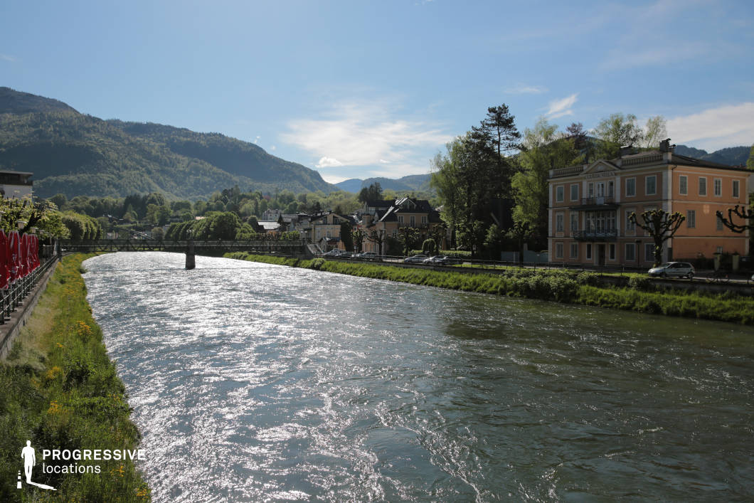 This location captures a gently curving river flanked by a low embankment and lined with manicured greenery, running through a quaint town nestled in a mountainous region. On the right, a three-story classical-style building with a muted peach façade and ornamental details stands prominently along the road, accompanied by geometrically pruned trees and bordered by a simple guardrail. The town's low-rise residential architecture blends traditional elements with a neutral color palette, harmonizing with the dense green hills and layered mountain ridges in the background. A narrow pedestrian bridge in the midground adds geometric interest and visual continuity across the waterway. The spatial composition is elongated and linear, creating a strong sense of depth and openness. This location is ideal for productions seeking a serene European riverside setting, suitable for drama, romance, or period pieces, and lends itself well to establishing shots, walking sequences, or tranquil transitional scenes.