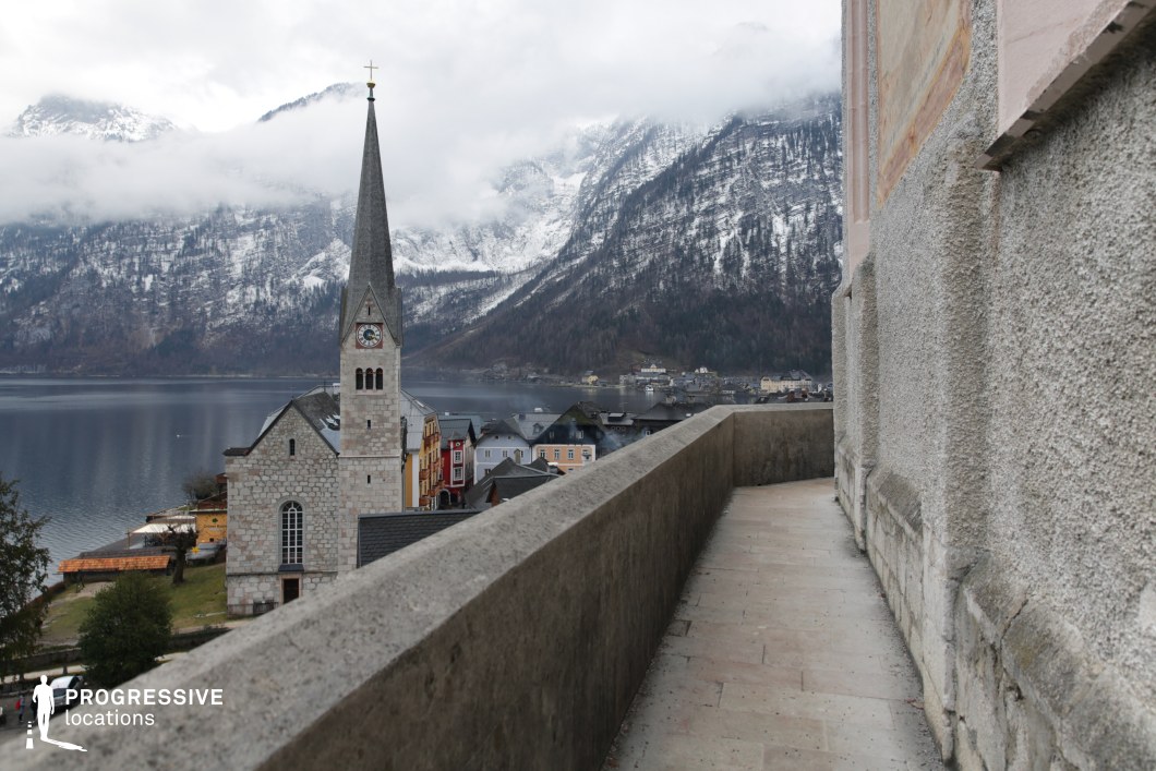 This image captures a narrow stone passageway running alongside a textured building wall, overlooking a lakeside town dominated by a prominent church with a tall, pointed spire and clock face. The foreground architecture features light-colored stone and rough plaster surfaces, while the town below presents a mix of pastel-toned façades in warm yellows, reds, and greys. Behind the church, a serene lake stretches across the frame, framed by dramatic snow-capped mountains with steep, heavily forested slopes, contributing to the depth and scale of the composition. The elevated viewpoint and clear visual layers—from foreground passage to midground architecture and background natural features—create a compelling sense of perspective. This location is ideal for filming period pieces, romantic dramas, or contemplative scenes, particularly those requiring a harmonious blend of historical architecture and majestic natural landscapes.