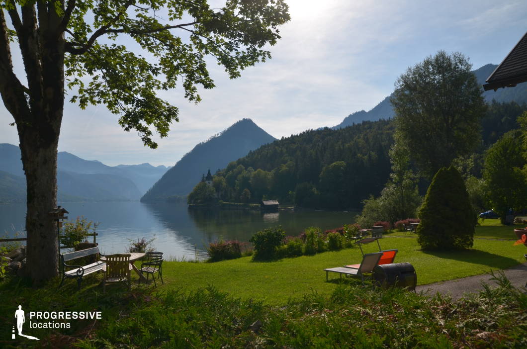 This serene lakeside location features a meticulously maintained grassy garden that gently slopes toward a still, expansive lake framed by densely forested hills and dramatic alpine peaks. In the foreground, benches, loungers, and a tree-shaded seating area create a tranquil, structured environment that contrasts with the rugged natural beauty in the background. The water's edge is softly bordered by flowerbeds and ornamental shrubs, while a small pointed-roof structure across the lake introduces a focal architectural element, harmoniously integrated into the tree-covered hillside. The composition offers a strong sense of depth, enhanced by layered mountain silhouettes and the reflective lake surface. Ideal for filming due to its quiet, undisturbed atmosphere, this setting is particularly suitable for genres such as drama, romance, and arthouse productions, and works well for contemplative scenes, introspective moments, or establishing shots of peaceful natural retreats.