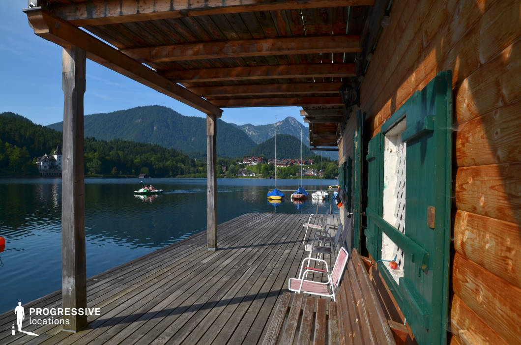 This image captures a spacious wooden terrace stretching along a calm lakeshore, with a pergola-style roof structure made from raw timber beams casting natural shadows onto the deck. The adjacent building features rustic log walls and green wooden shutters, enhancing the traditional alpine character. The open planked flooring extends seamlessly to the water’s edge, where small sailboats are moored, and the opposite side of the lake is framed by forested hills and alpine peaks, creating a harmonious natural backdrop. The symmetry of the terrace beams and the perspective leading into the mountainous horizon offers a strong geometric composition ideal for framing cinematic shots. This lakeside location is highly suitable for filming due to its picturesque scenery, architectural authenticity, and unobstructed sightlines—perfect for drama, romance, travel-themed productions, or tranquil lifestyle scenes.
