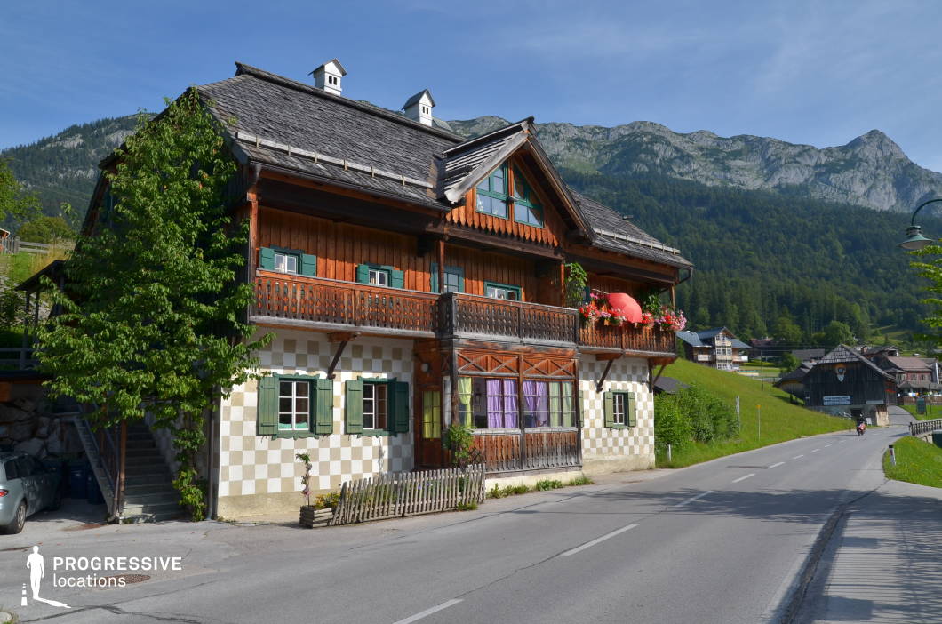 This location features a traditional alpine-style house situated directly beside a gently curving asphalt road, set against a mountainous forested backdrop. The two-story building combines a distinctive checkerboard-patterned ground floor with green shuttered windows and an upper level clad in warm-toned timber, adorned with carved wooden balcony railings and decorative gables. A small enclosed veranda with large paneled windows and vivid curtains adds texture and contrast to the façade. The grey-tiled pitched roof is steep and detailed with dormers, contributing to the structure’s rustic character. The surrounding environment includes open grassy inclines, additional wooden structures, and dramatic rocky hills rising in the distance. This visually cohesive and serene rural setting is ideal for filming scenes requiring an authentic alpine village atmosphere. Suitable for genres such as drama, period films, or romantic stories, the location works well for establishing shots, exterior dialogues, or transitional sequences in natural or small-town narratives.