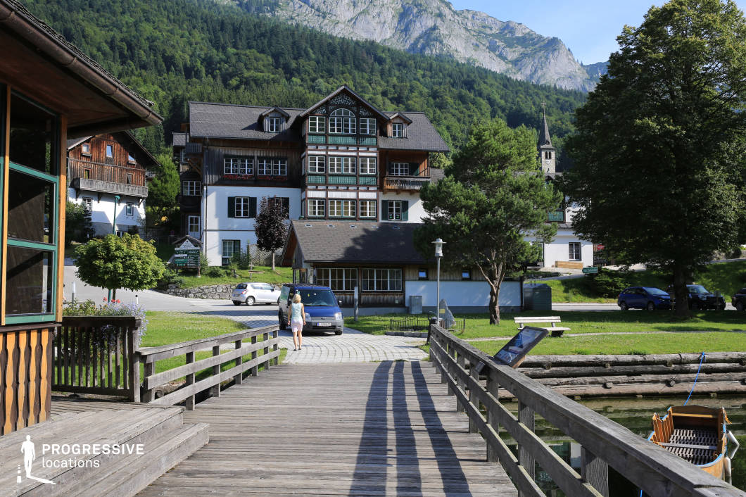 This location features a traditionally styled alpine building nestled at the base of a densely forested mountain slope. The architecture showcases a combination of whitewashed stone and dark wooden cladding, with symmetrical window arrangements and intricate gable detailing that highlight its regional character. In the foreground, a wide wooden boardwalk with evenly spaced railings leads from a lakeside dock area toward the building, visually connecting water and settlement. The setting is structured with a variety of vertical and horizontal lines — from the planks of the dock to the multi-level rooflines — creating depth and compositional balance. Surrounding greenery and mature trees provide natural framing, while the backdrop of rugged, rocky peaks enhances the dramatic scale. This location is ideal for filming serene rural scenes, character-driven dramas, or period pieces, offering a harmonious blend of built heritage and natural grandeur.