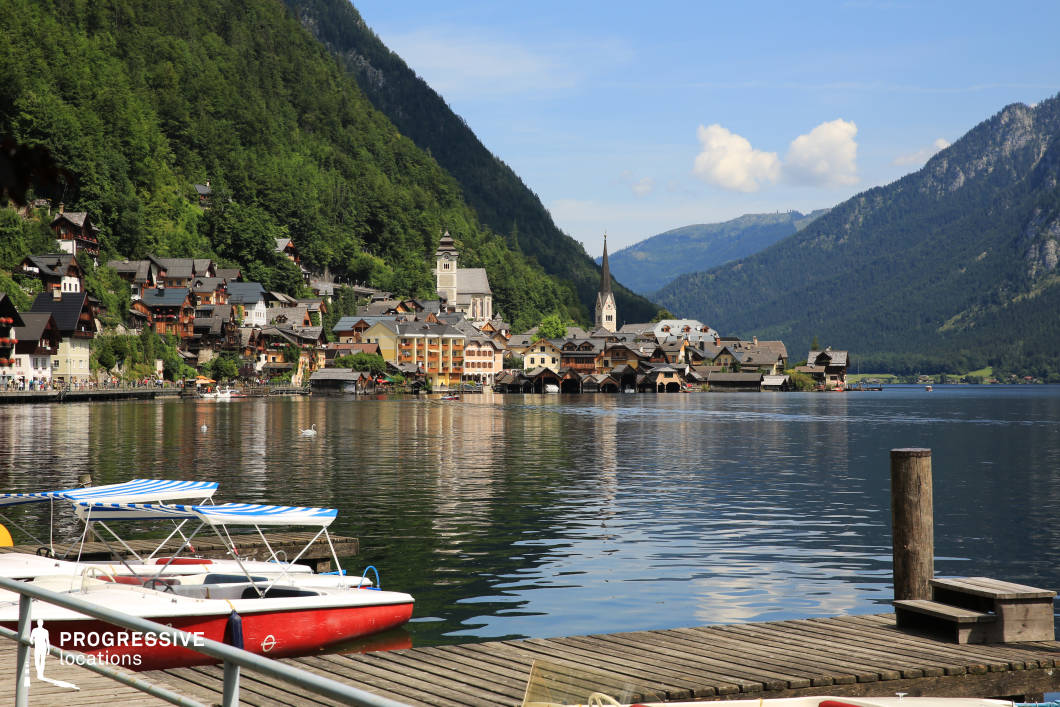 This lakeside village scene features a harmonious blend of traditional Alpine architecture nestled at the base of steep, forest-covered mountains. Dominated by gabled roofs and pastel-toned facades, the buildings cluster tightly along the water's edge, anchored visually by a tall, slender church spire and a more robust bell tower—both serving as central architectural anchors. The water in the foreground is calm and expansive, with a wooden pier and moored boats providing a functional, accessible element to the scene. The composition is framed by high, green mountain slopes and distant ridges, creating a strong sense of enclosure and depth. The color palette is natural and cohesive, with rich greens, soft earth tones, and neutral greys that support a peaceful, authentic alpine ambiance. This location is ideal for filming serene lakeside settings, romantic or historical genres, and scenes requiring a picturesque European village backdrop.
