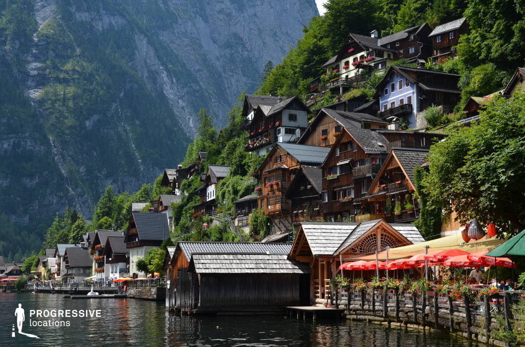 This image captures a densely layered hillside village composed of traditional alpine-style buildings, nestled between a steep, forested mountain backdrop and a serene lakeside. The architecture features steep gabled roofs, exposed wooden façades, and vibrant flower boxes, creating a visually cohesive and rustic aesthetic. The buildings are arranged in a cascading formation, giving the composition a strong vertical rhythm and sense of depth. The dominant materials include wood and stone, reinforcing a natural, handcrafted feel, while the rich greenery surrounding the structures further integrates the village into its mountainous environment. At the waterfront, boathouses and a wooden promenade with vibrant red parasols introduce a functional leisure space, creating a contrast between the vertical hillside density and the horizontal calm of the lake. This location is highly suitable for filming due to its immersive, storybook-like atmosphere and strong visual character, making it ideal for genres such as drama, historical fiction, fantasy, or travel-related productions, and particularly effective for establishing shots, scenic transitions, or quiet reflective scenes.