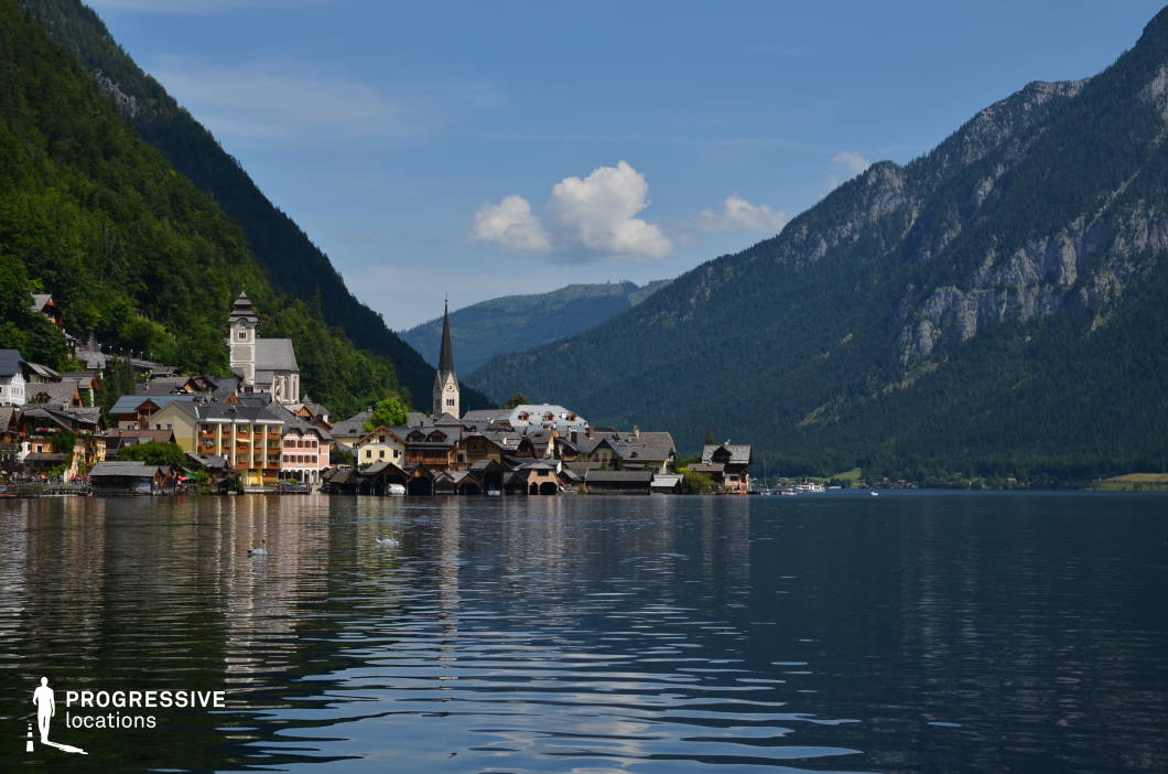 This panoramic location presents a compact lakeside village nestled between steep, forested mountains and a calm, reflective body of water. The architecture is characterized by a blend of traditional alpine elements—pitched roofs, wooden balconies, pastel-colored facades—and distinctive landmarks, such as a central church with a tall, pointed steeple and a historic-looking stone tower. The visual rhythm is dominated by the contrast between the dark green slopes and the smooth surface of the lake, which amplifies the scene’s symmetry and balance. The setting is visually enclosed yet expansive, with the surrounding natural walls of rock and tree-covered elevations adding depth and drama. The harmonious integration of human-made and natural elements creates a timeless, serene atmosphere. This location is particularly suitable for historical, romantic, or dramatic genres, and works well for establishing shots, introspective scenes, or any visual storytelling that benefits from a picturesque, secluded alpine environment.