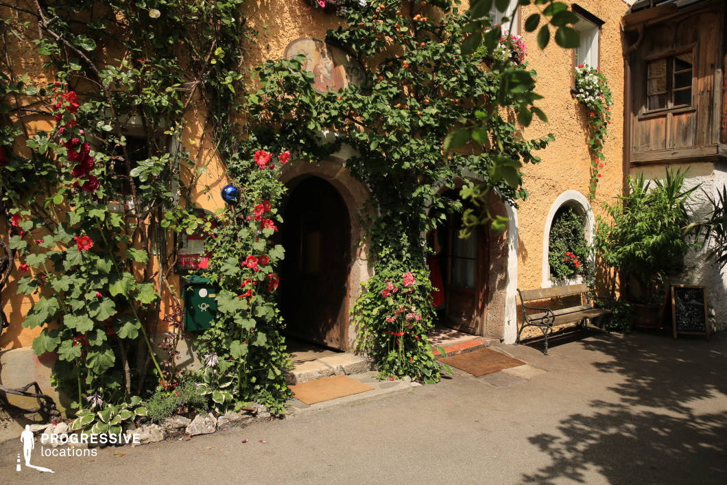 This charming location features the entrance of a traditional plastered building with a warm, textured ochre façade partially clad in climbing ivy and vibrant red flowering plants. The arched wooden door is framed by natural stone, giving a historic and slightly worn appearance, while small architectural details like a half-timbered window bay and an arched white-trimmed window add depth and character. A wooden bench and potted greenery further enrich the scene, contributing to a serene, intimate atmosphere. The composition is well-balanced with a narrow paved forecourt allowing close-range filming or photography. The visual harmony of natural textures—plaster, stone, and wood—along with the rich greenery and saturated colors, creates a timeless, European small-town aesthetic. This location is ideal for period dramas, romantic scenes, or character-driven stories requiring a rustic and inviting setting.