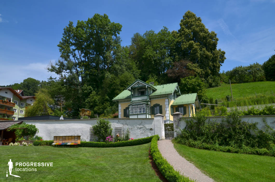 This location features a charming, two-story villa with classical architectural details and a green gabled roof that contrasts harmoniously with its pale yellow facade and dark green window shutters. The structure is set behind a white perimeter wall with a wrought iron gate, accessible via a narrow gravel pathway flanked by neatly trimmed hedges. The composition is defined by the villa’s symmetry, wooden balcony details, and central placement within a lush, elevated landscape backed by dense mature trees and open grassy slopes. The foreground lawn is expansive and well-maintained, offering visual clarity and depth. The setting balances built and natural elements, ideal for productions requiring a secluded, picturesque residential estate. Its timeless aesthetic and layered perspectives make it suitable for drama, historical pieces, or elegant commercial scenes.