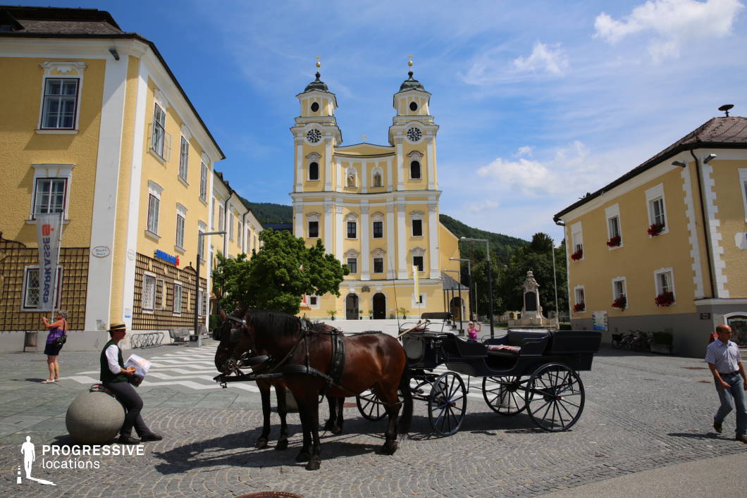 This photograph captures a spacious, visually balanced town square dominated by a grand baroque church with twin bell towers and a richly articulated yellow-and-white façade. The surrounding buildings echo the church's color palette, creating a cohesive and harmonious streetscape. The structures are two to three stories high with traditional gabled roofs and uniformly arranged windows, contributing to a symmetrical and orderly visual rhythm. The square itself is paved with cobblestone, adding historical texture and authenticity. Geometrically, the composition is defined by axial symmetry centered on the church, with linear perspectives leading the eye toward the entrance. The location is visually open, with a slight elevation change at the church entrance that adds depth. Ideal for filming due to its strong architectural character, the square is suitable for period pieces, historical dramas, romantic scenes, or European cityscapes requiring a classical and elegant atmosphere.