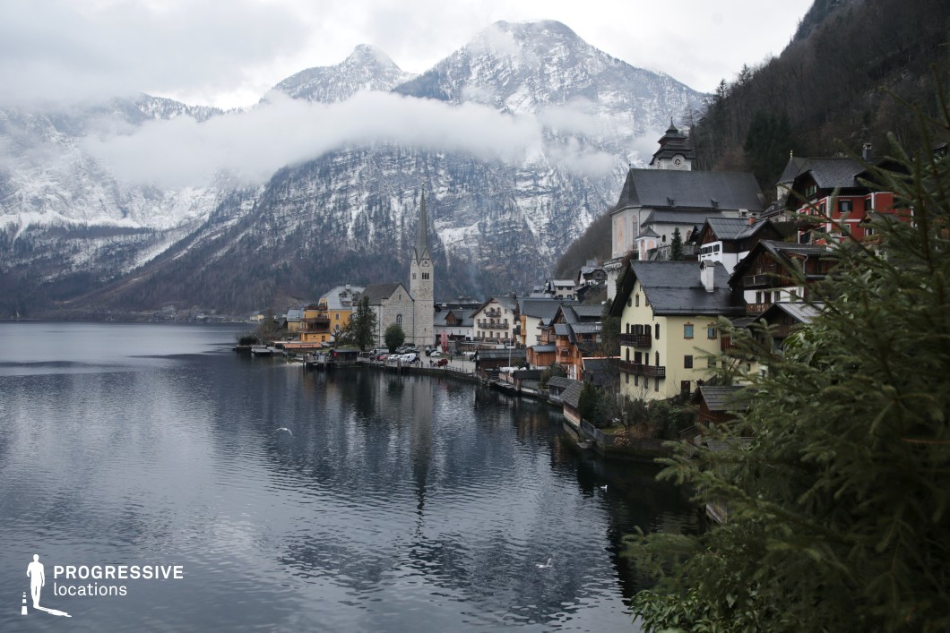 This location features a compact, picturesque lakeside village nestled against a dramatic alpine backdrop. Dominated by steep, snow-dusted mountain slopes and a tranquil body of water, the setting includes traditional multi-story buildings with gabled roofs, muted pastel facades, and wooden balconies that climb the hillside in layered rows. A tall, narrow church steeple constructed from grey stone provides a central vertical focal point, complemented by the substantial presence of a hilltop church complex. The built environment exhibits classic alpine architectural elements, with slate roofs and timber accents enhancing the setting's historic and regional character. The clear spatial hierarchy, from the lakeshore structures to the elevated religious buildings, provides a strong sense of depth and scale. The location is ideal for filming due to its cohesive visual identity and immersive natural setting, making it particularly suitable for historical dramas, romantic films, or any narrative requiring an atmospheric European village or mountainous lakeside environment.