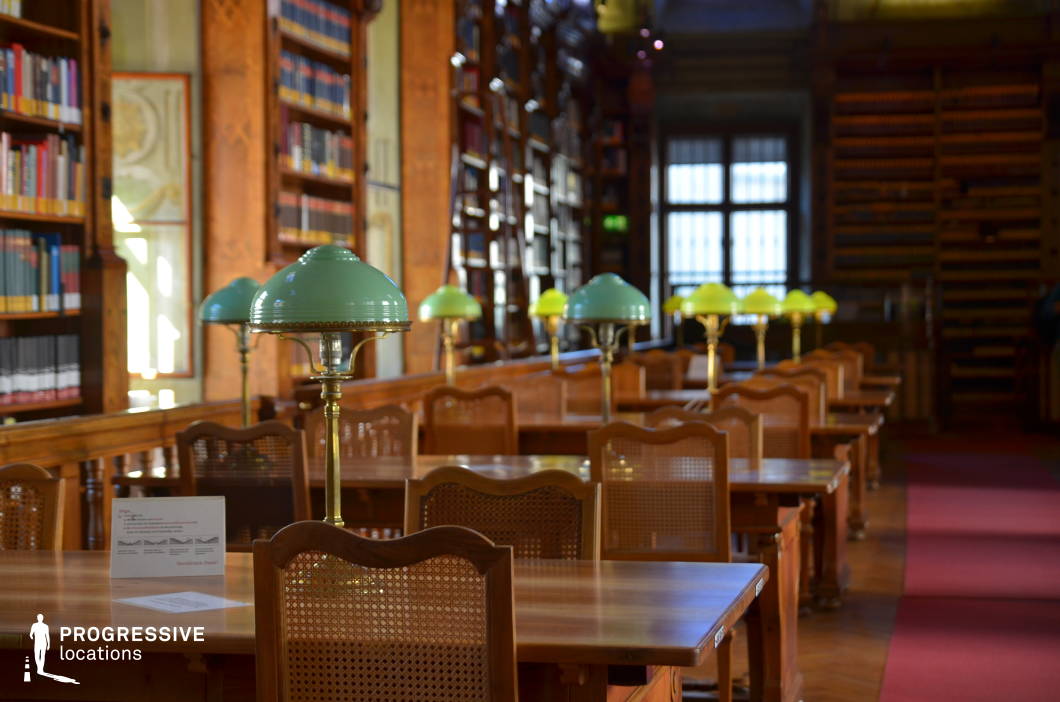 This elegant reading room exudes a timeless academic atmosphere, featuring long wooden tables arranged in neat rows, each adorned with vintage green-shaded brass lamps. The warm wood tones of the bookshelves, lined with neatly arranged volumes, contribute to the refined aesthetic. Tall windows framed by dark wood allow soft, diffused light to filter in, enhancing the tranquil and scholarly ambiance. The red carpeted walkway adds a sense of prestige and structure to the space. This location is ideal for filming academic dramas, period films, intellectual thrillers, or introspective character moments, providing a visually rich and historically evocative setting.