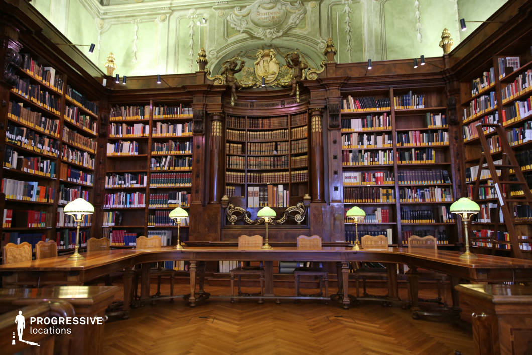 This image captures a grand historic library reading room with rich wooden bookshelves extending to the ceiling, filled with neatly arranged volumes. The space is accentuated by Baroque architectural details, including gilded ornamentation, sculpted reliefs, and an elegant curved bookshelf at the center. The pale green walls with intricate stucco work contrast beautifully with the dark wood, enhancing the refined aesthetic. A long wooden table with classic reading lamps featuring green glass shades reinforces the scholarly ambiance. This setting is ideal for filming period dramas, mystery thrillers, and academic or legal narratives, offering a visually immersive backdrop for scenes requiring an atmosphere of knowledge, prestige, or intellectual intrigue.