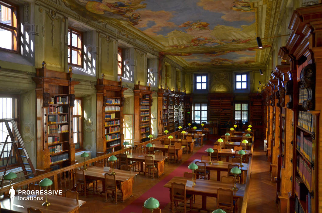 This opulent historic library features towering wooden bookshelves with intricate carvings, filled with an extensive collection of books. The room is defined by its high, elaborately decorated ceiling adorned with classical fresco paintings, lending a sense of grandeur and artistic depth. Tall arched windows allow natural light to filter in, casting soft shadows on the detailed architectural moldings along the walls. The long wooden tables, each equipped with vintage green-shaded reading lamps, create a scholarly atmosphere. The rich wood tones of the furniture and flooring contrast with the muted green hues of the walls, evoking a timeless elegance. This location is ideal for filming period dramas, academic thrillers, or historical documentaries, offering an authentic intellectual and aristocratic ambiance suitable for scenes set in old universities, secret societies, or scholarly research environments.