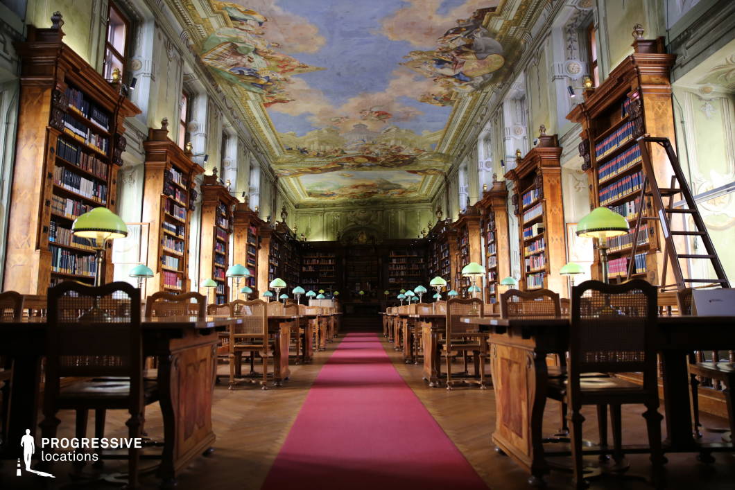 This opulent library hall exudes a rich, classical ambiance with its towering wooden bookshelves, intricate architectural details, and a stunningly painted ceiling depicting baroque-style frescoes. The symmetrical rows of wooden reading desks, adorned with vintage green-shaded lamps, create a refined and structured aesthetic, further emphasized by a long red carpet running down the center. Large windows allow natural light to filter in, complementing the warm tones of the wood and the elaborate wall ornamentation. The high ceiling and grand proportions of the space enhance its sense of prestige and historical significance. This location is ideal for filming historical dramas, intellectual thrillers, or period-piece settings requiring an academic or scholarly atmosphere. The elegant symmetry and rich textures also make it well-suited for refined, contemplative, or high-stakes intellectual exchanges.