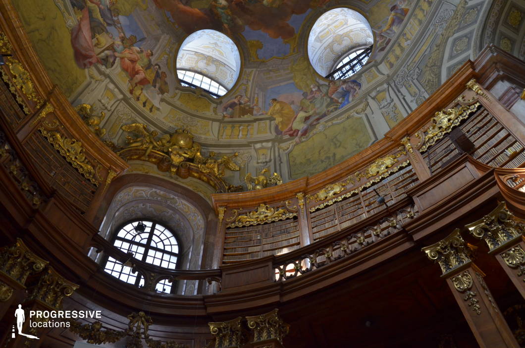 This grand Baroque library features a soaring domed ceiling adorned with elaborate frescoes depicting classical allegories in vibrant hues, framed by intricate stucco work and gold accents. The circular upper gallery, constructed of dark polished wood with gilded carvings, houses an extensive collection of meticulously arranged books, emphasizing the intellectual grandeur of the space. Large arched windows punctuate the curved walls, allowing natural light to filter through, highlighting the interplay of light and shadow on the richly decorated architectural details. The space is ideal for filming historical dramas, academic thrillers, or fantasy settings that require a regal, scholarly, or mysterious atmosphere.