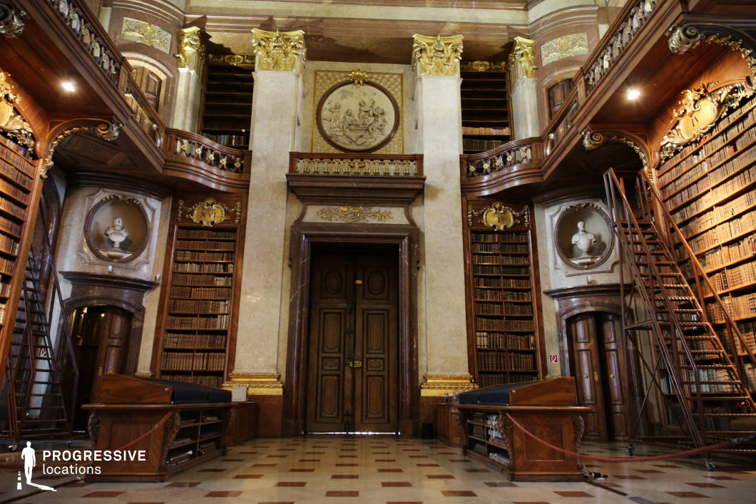 This opulent library hall exudes classical grandeur, featuring towering bookshelves, rich wooden paneling, and elaborate gold embellishments. A central double door, framed by marble columns and an ornate pediment, serves as a focal point, enhancing the sense of formality and prestige. Above, a circular relief and intricate gilded details reinforce the room’s historical and intellectual significance. Curved balconies with decorative railings wrap around the space, allowing access to upper-level bookshelves via elegant wooden ladders. The polished checkered floor reflects the warm glow of the ambient lighting, creating an inviting yet imposing atmosphere. This location is ideal for filming period dramas, scholarly intrigue, or regal settings, offering a distinguished backdrop for academic debates, secret discoveries, or aristocratic storytelling.