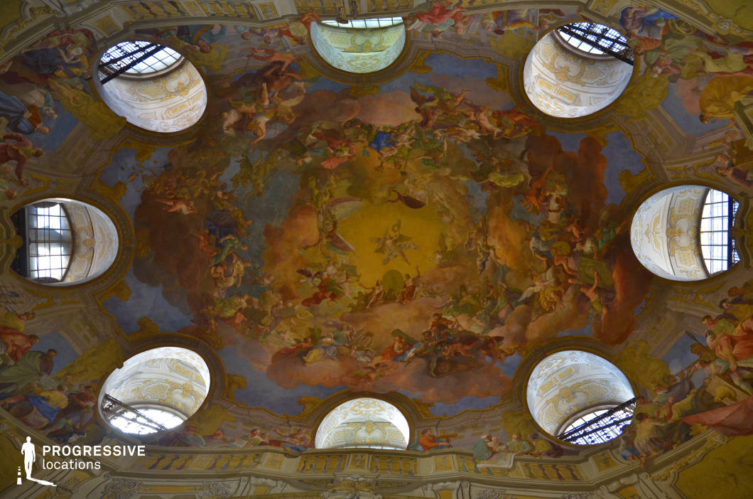 The image captures an opulent Baroque-style ceiling fresco, rich in vibrant colors and dynamic compositions. The artwork, set within a domed architectural space, depicts a celestial scene filled with figures floating among clouds, radiating from a central golden focal point. The fresco is framed by ornately decorated moldings and large, symmetrically arranged circular windows that allow natural light to highlight the intricate details. The grandeur of this space makes it ideal for filming historical dramas, fantasy epics, and period pieces, offering an authentic setting for scenes requiring a sense of divine majesty, aristocratic refinement, or artistic brilliance.