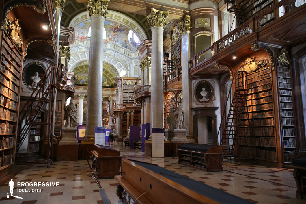 This opulent library showcases intricate Baroque architecture, featuring towering bookshelves adorned with gilded carvings, marble columns, and a grand frescoed ceiling. The symmetrical layout is accentuated by wooden balconies with ornate railings, accessible by elegant staircases. Busts and statues integrated into the design add a sense of historical grandeur. The checkered marble flooring enhances the classical aesthetic, reflecting the warm tones of the wood and gold detailing. The interplay of natural and artificial lighting highlights the rich textures and depth of the space. This setting is ideal for period dramas, intellectual thrillers, or historical narratives, offering a visually stunning backdrop for scenes involving scholars, secret discoveries, or intellectual debates.
