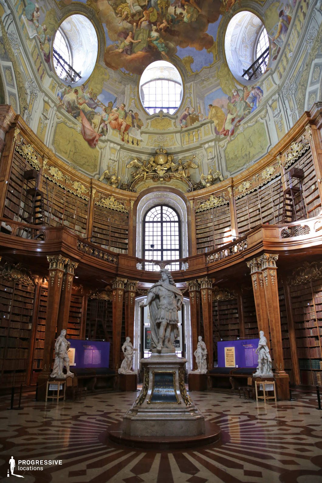 This opulent Baroque-style library features towering wooden bookshelves adorned with intricate gold detailing, creating a sense of grandeur and historical depth. A central marble statue stands as the focal point, surrounded by additional sculptural elements that enhance the room’s regal atmosphere. The domed ceiling is lavishly decorated with frescoes, featuring dramatic scenes in soft pastel hues, framed by elegant circular windows that allow natural light to filter in. The symmetrical composition and intricate detailing make this location ideal for filming historical dramas, period pieces, or fantasy films requiring an aristocratic or scholarly ambiance. Scenes involving secret knowledge, intellectual discourse, or mystical discoveries would be especially well-suited to this setting.