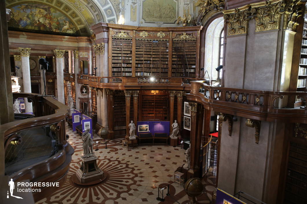 This grand library hall showcases an opulent Baroque design with towering wooden bookshelves adorned with gold embellishments, supporting an extensive collection of antique volumes. The high, frescoed ceilings feature dramatic allegorical scenes, further enhancing the room’s historical grandeur. The space is structured around a central circular floor with an intricate geometric tile pattern, surrounded by imposing marble columns and sculpted statues that add a classical elegance. An upper gallery with a carved wooden balustrade provides a striking perspective, emphasizing the library’s scale and symmetry. This location is ideal for filming period dramas, historical thrillers, or scholarly-themed narratives, offering a setting of intellectual sophistication and aristocratic refinement.