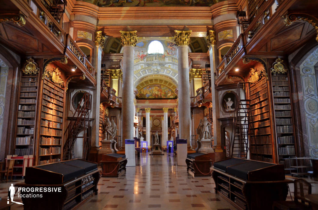 The image showcases a majestic Baroque-style library hall with towering bookshelves reaching up to an ornately decorated mezzanine level. Intricately carved wooden balustrades, gold-accented architectural details, and classical sculptures contribute to the grandeur of the space. The ceiling is adorned with frescoes depicting mythological or historical scenes, framed by massive marble columns with gilded capitals, reinforcing the opulent aesthetic. The symmetrical layout and polished checkered floor enhance the sense of depth and perspective. This location is ideal for filming historical dramas, period pieces, and intellectual thrillers, as well as scenes requiring a setting of scholarly elegance, aristocratic refinement, or secretive, knowledge-laden intrigue.