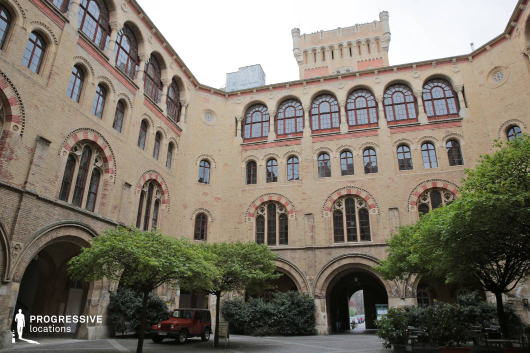 This enclosed courtyard is surrounded by a grand historic brick building featuring arched windows, decorative stonework, and a fortified central tower. The Romanesque and neo-Gothic architectural elements, such as rounded archways and ornamental red brick details, create a sense of historical depth. The open courtyard, framed by lush greenery and symmetrical facades, adds a balanced composition to the space. The arched entranceway leads into the structure, enhancing the cinematic potential for dramatic entrances or transitions. This location is ideal for historical films, medieval-inspired narratives, or period dramas, offering a stately and fortress-like ambiance suited for institutional, academic, or military-themed productions.